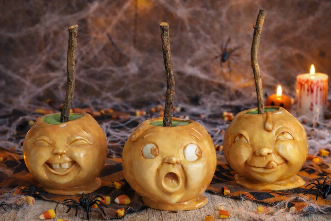 Three caramel-coated toffee apples with expressive faces, displayed in a Halloween scene with candy corn, spiders, and candles.