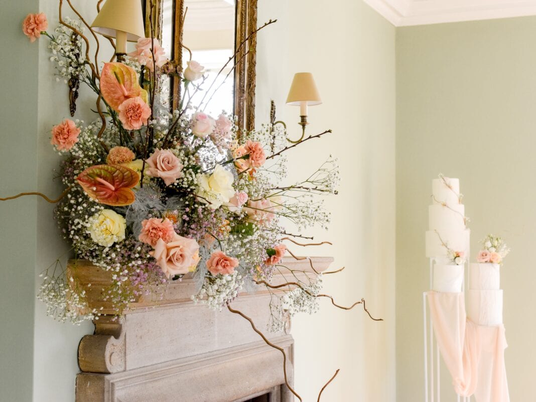 Elegant wedding venue with a lush floral mantel arrangement in soft pink and white blooms, with a tiered wedding cake in the background.