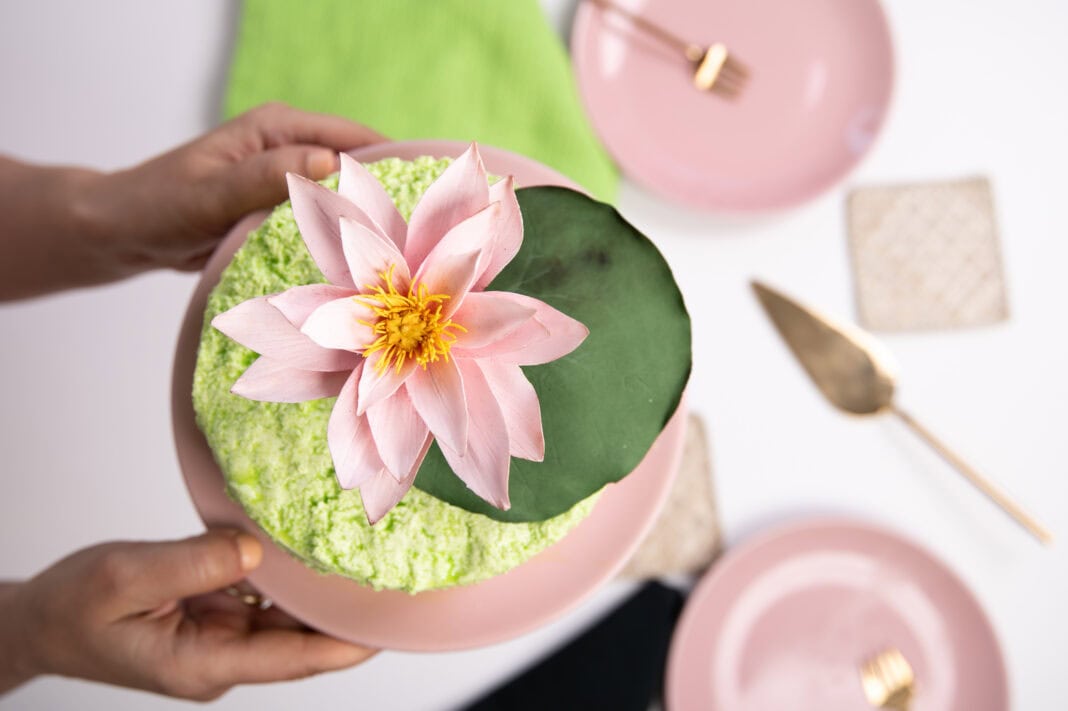 Green cake topped with a pink gum paste water lily flower and a lily pad, photographed from above.