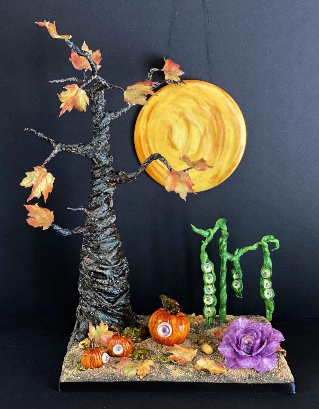 Halloween-themed sugar art display featuring a twisted tree, pumpkins with eyeballs, pea pods with eyes, autumn leaves, and a large full moon.