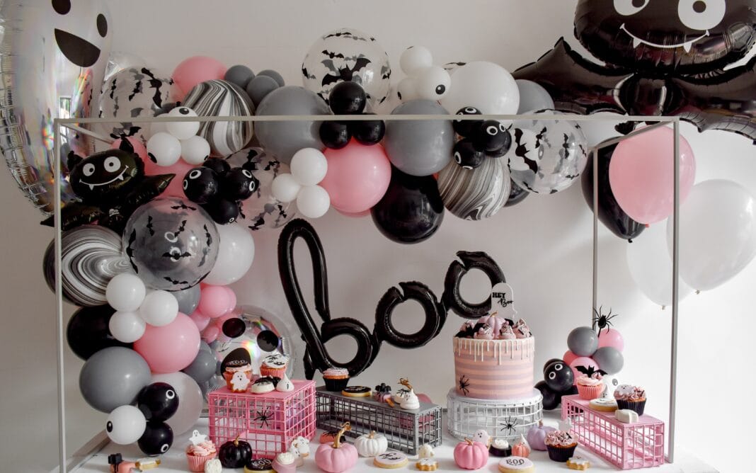 Pink Halloween dessert table decorated with black, white, gray, and pink balloon garlands, a Halloween cake, cupcakes, cookies, and pumpkin-themed treats.