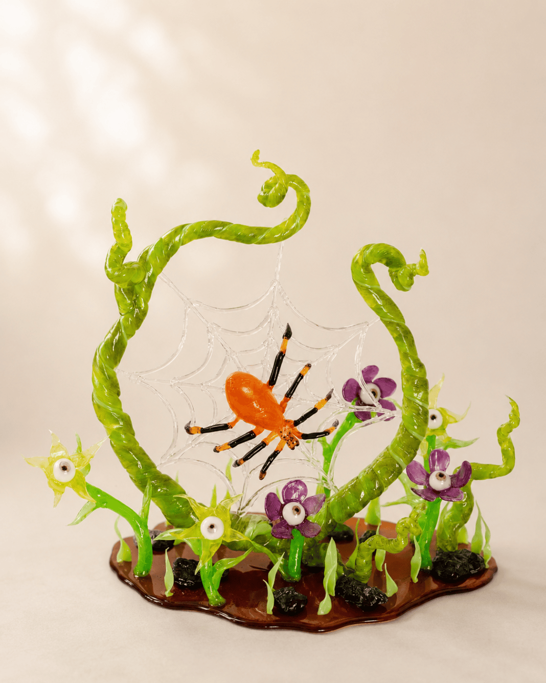 Intricate isomalt display featuring a bright orange spider suspended in a clear sugar web surrounded by twisting green vines and whimsical flowers with eyeball centers. The sculpted sugar arrangement sits on a glossy amber base with small leaves and dark stones, creating a playful Halloween-inspired centerpiece against a soft neutral background.