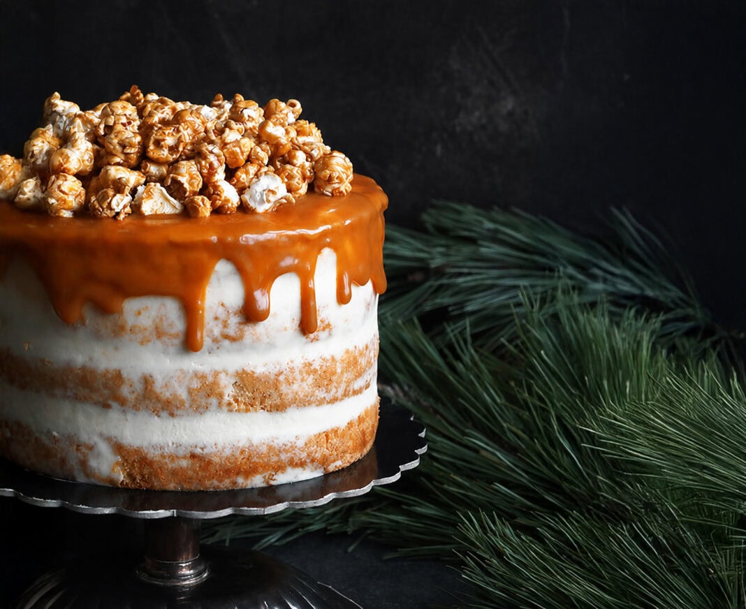 Caramel Apple Popcorn Cake with layers of vanilla cake and frosting, topped with a glossy caramel drip and a mound of caramel popcorn against a dark rustic background.