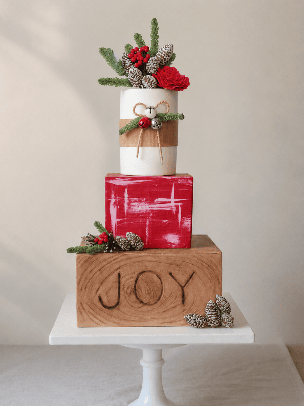 A festive three-tier Christmas-themed cake sits on a white pedestal stand against a soft neutral background. The bottom tier resembles a carved wooden block with the word “JOY,” topped with a red gift-inspired tier and a white cylindrical tier decorated with ribbon, greenery, and jingle bells. Pinecones, evergreen sprigs, red berries, and a red flower adorn the top and sides, creating a rustic holiday centerpiece.