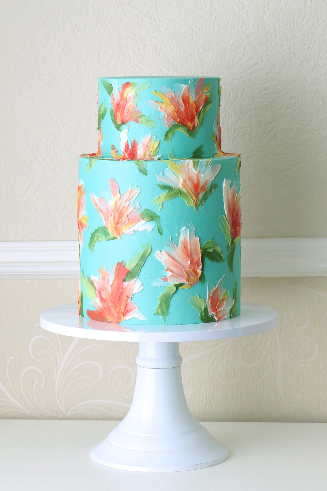 Two-tier turquoise buttercream cake decorated with hand-painted coral, peach, and white floral brushstrokes with green leaves, displayed on a white cake stand against a soft neutral background.