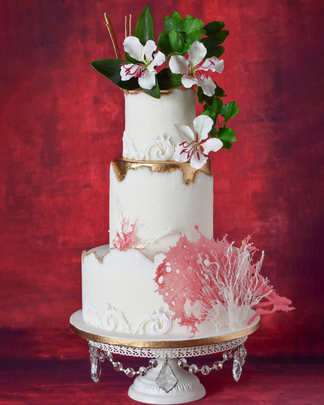 Three-tier white wedding cake with textured buttercream, gold accents, delicate piped scrollwork, pink coral-inspired sugar decorations, and tropical white flowers with green foliage cascading from the top, displayed on an ornate crystal cake stand.