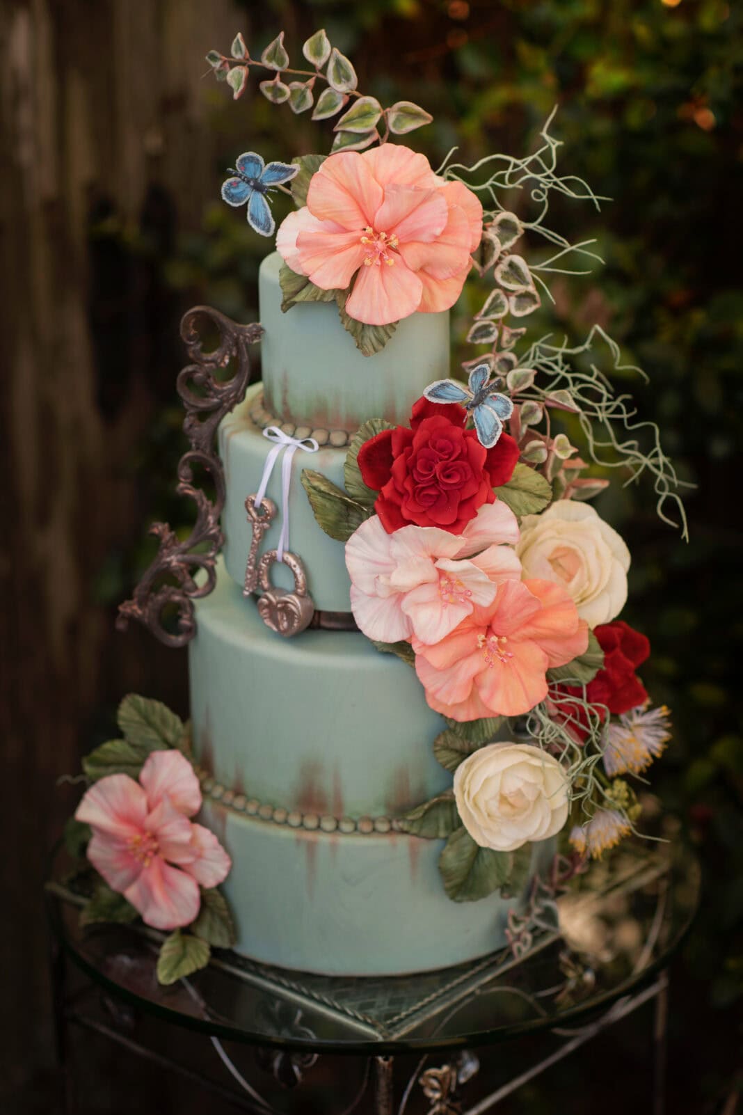 A three-tier pastel sage-green fondant cake decorated with intricate handmade sugar flowers, including large peach-pink hibiscus-style blooms, red roses, cream roses, green leaves, and small blue flowers. Delicate trailing vines and pale green tendrils extend from the floral arrangements. The cake features antique bronze decorative accents, including ornate scrollwork, a ribbon-like embellishment, and beaded borders between the tiers. A large floral arrangement cascades diagonally down the right side, with an additional pink flower on the lower left. The cake is photographed outdoors against a softly blurred, dark green garden background.