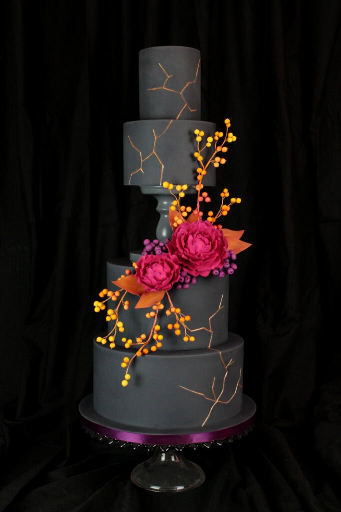 A tall, dark gray tiered cake decorated with metallic gold crack details, vibrant pink peonies, purple berries, orange leaves, and cascading orange and yellow berry branches against a black backdrop.
