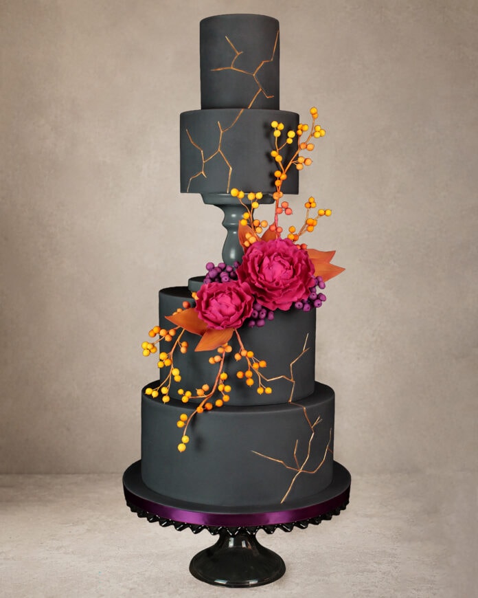 A tall, dark gray tiered cake decorated with metallic gold crack details, vibrant pink peonies, purple berries, orange leaves, and cascading orange and yellow berry branches against a black backdrop.